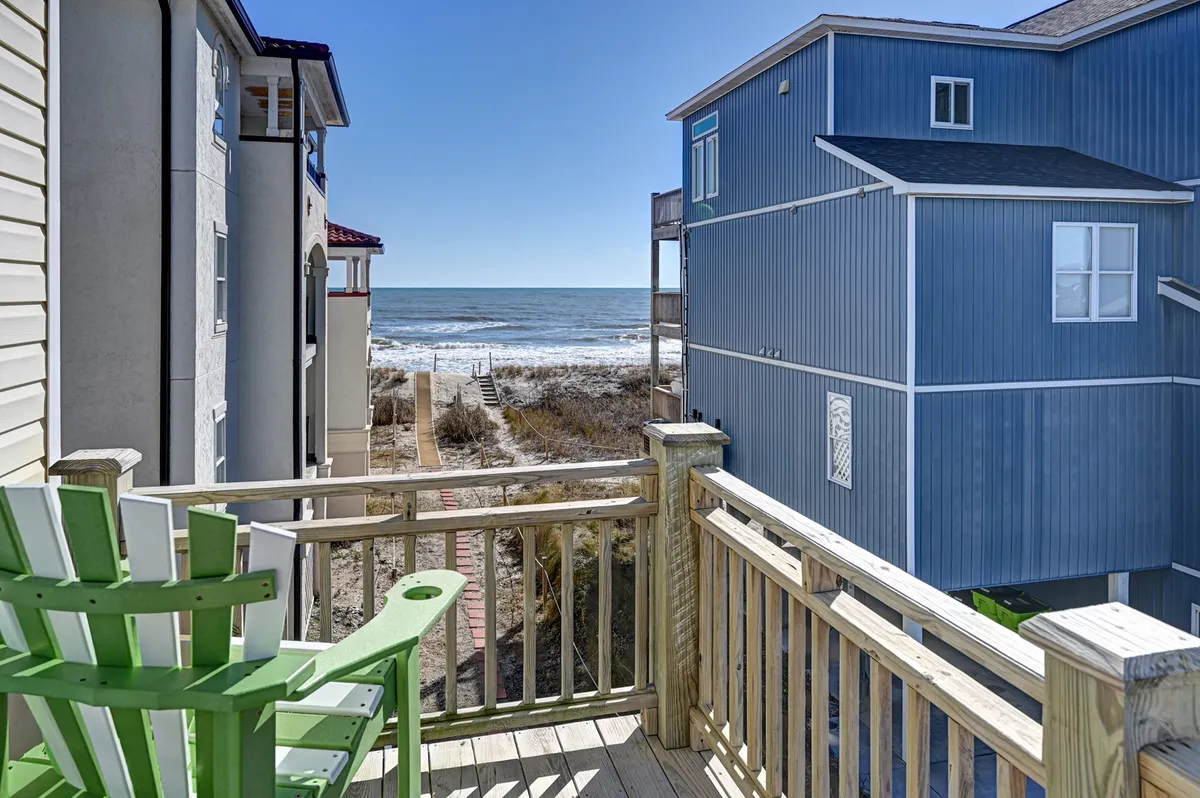 A tranquil wooden deck outfitted with a green Adirondack chair, offering unobstructed ocean views beyond the railing. Neighboring beach houses dot the landscape, and the deck is bathed in sunlight—inviting relaxation and family gatherings outdoors.