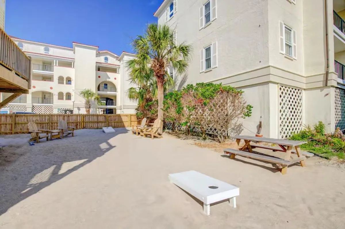 A vibrant backyard scene featuring a picnic table set beneath the shade of palm trees, a cornhole game on a sand area, and open space for outdoor fun. The mood is casual and inviting, with plenty of room for both play and relaxation, making it ideal for gatherings and family activities.