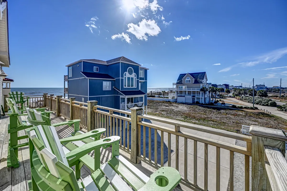 This exterior image showcases green Adirondack chairs positioned perfectly on a spacious deck, looking out over a stunning ocean view with neighboring houses visible in the distance. The scene is bathed in natural light, capturing a casual, welcoming atmosphere ideal for relaxation or socializing.