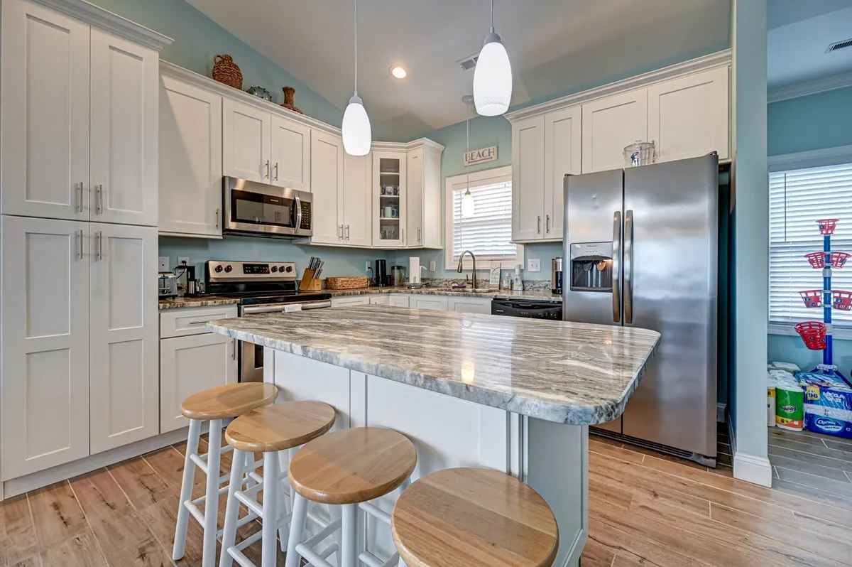 This kitchen-focused image highlights a granite countertop with stools for island seating, stainless steel appliances, and warm natural light streaming through the window. The open kitchen is inviting and clearly designed for both cooking and gathering, with an airy, coastal feel.