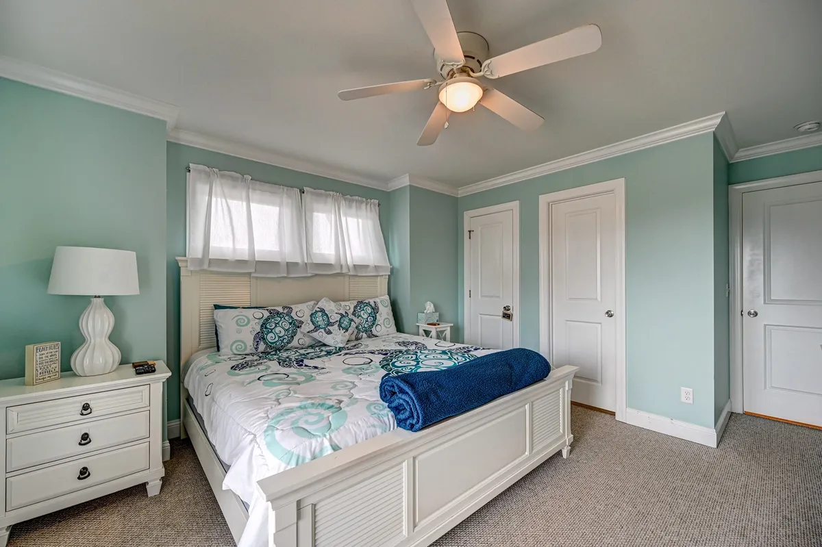 This king bedroom is filled with natural light from large windows draped in sheer curtains. The room features a bed with patterned bedding, a white nightstand, and a ceiling fan, creating a cozy, inviting atmosphere that feels both relaxing and luxurious—perfect for highlighting the comfort and privacy described in the section.