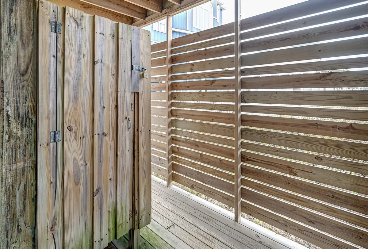 A close-up view of a wooden outdoor shower stall surrounded by slatted fencing, sunlight filtering through. The scene suggests a refreshing place to rinse off after a day at the beach or playing outdoors, contributing to the sense of easy, seamless transitions between fun and relaxation.
