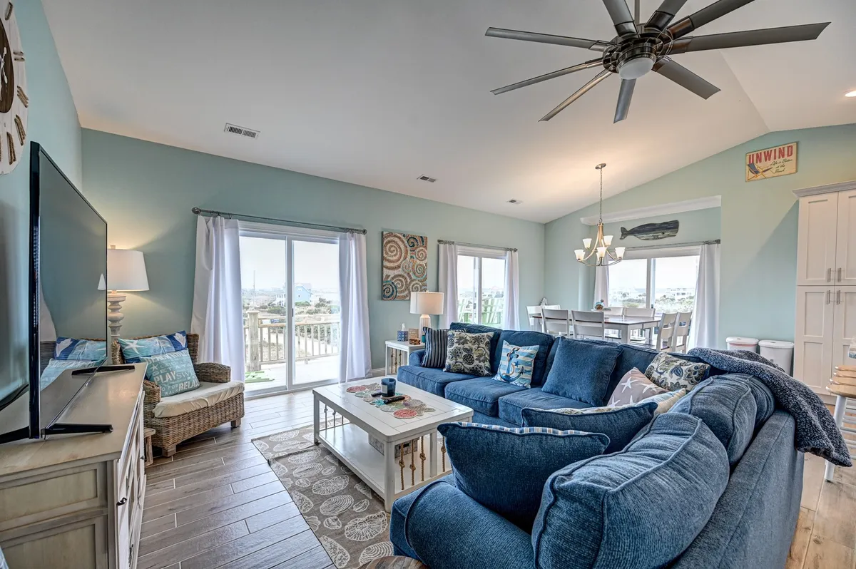 A welcoming interior scene featuring a spacious blue sectional sofa, a large flat-screen TV, a dining area with a view, and stylish coastal decor. The arrangement invites both cozy movie nights and lively gatherings, with elements of comfort and relaxation at the forefront.