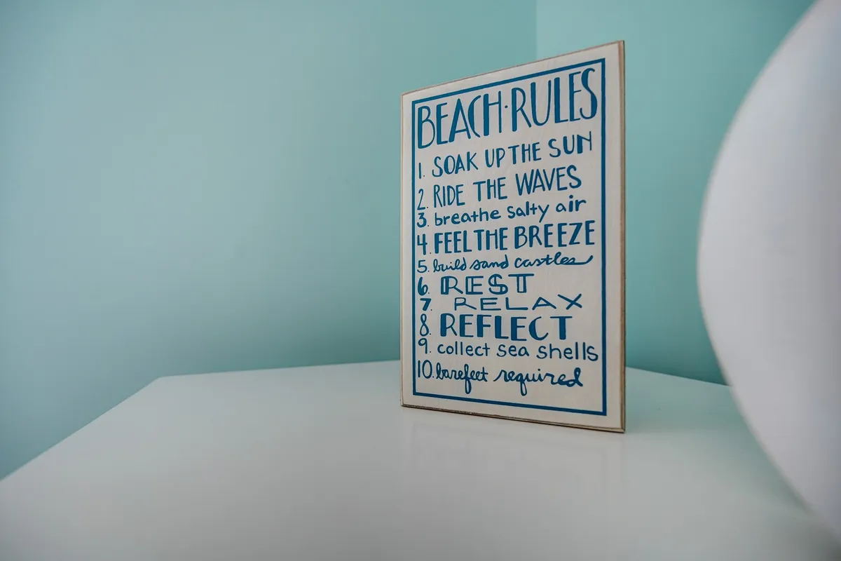 A playful interior scene with a 'Beach rules' sign set against a light-colored table and soft blue wall. The decor invokes themes of fun, sun, and family, referencing beach traditions and local adventures.