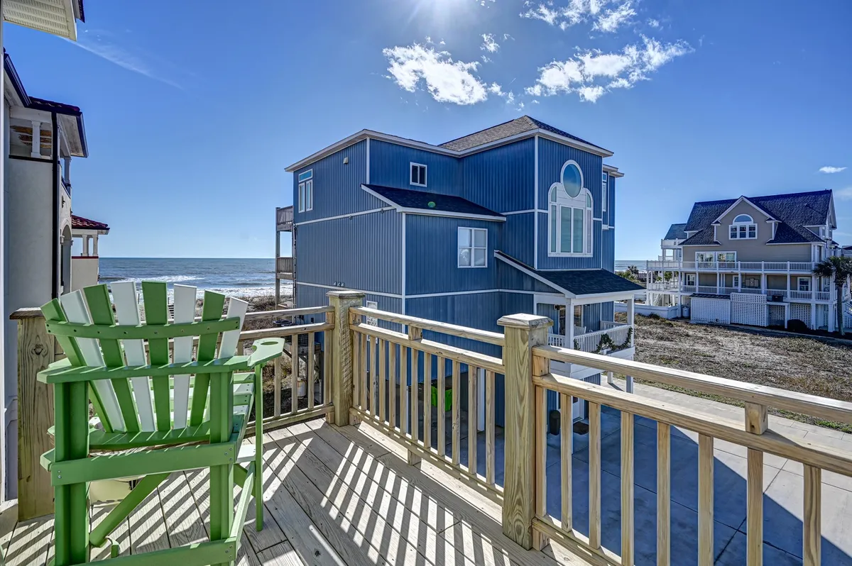 An inviting exterior scene featuring a green adirondack chair placed on a deck overlooking a sandy beach and blue house with a wide sky above. The tranquil setting directly connects the home to the natural beauty and recreational opportunities of the coastline.