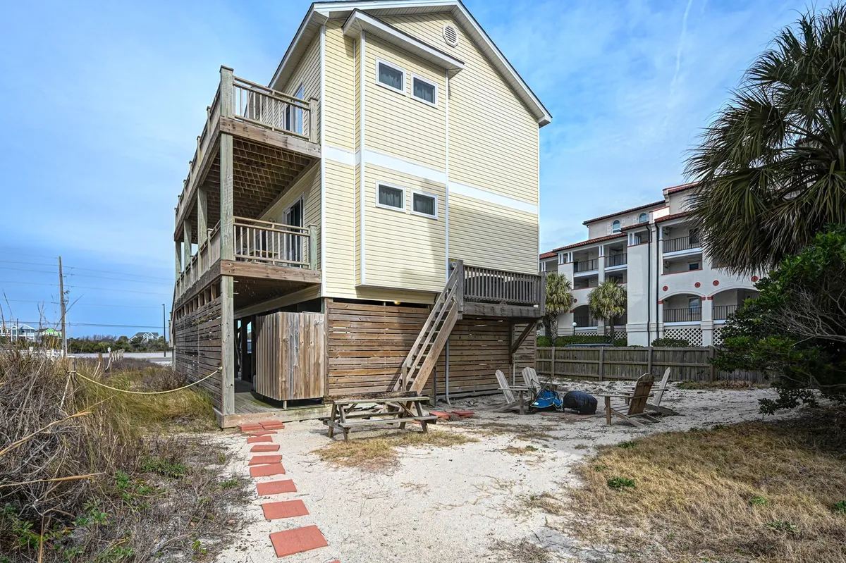 A wide wooden deck leads to a sandy pathway surrounded by palm trees, blending the comforts of the home with direct outdoor access. The setting is perfect for sunbathing, morning coffee, or gathering before heading to the beach.