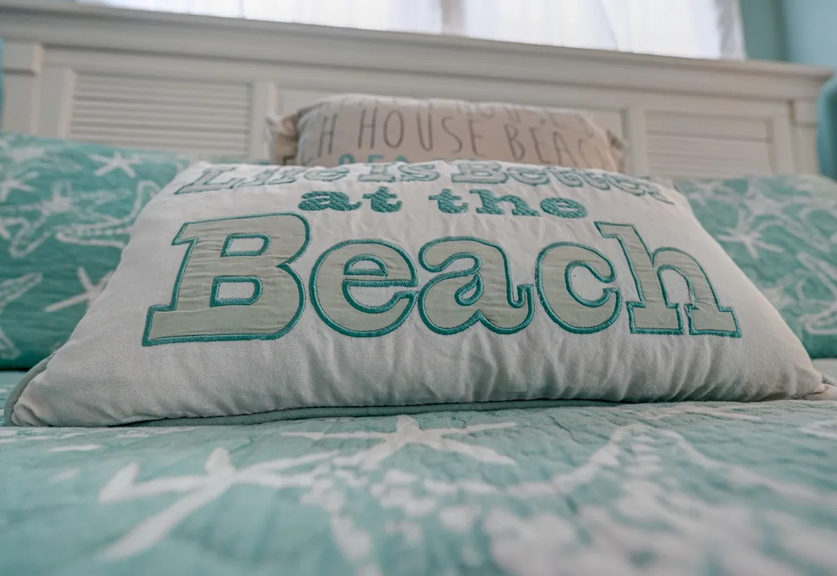 A cheerful bedroom scene with a decorative pillow featuring text, a beach-themed quilt, and a light-colored bed frame, all bathed in natural light. The space feels cozy and meticulously prepared, reflecting both relaxation and attention to guest comfort.