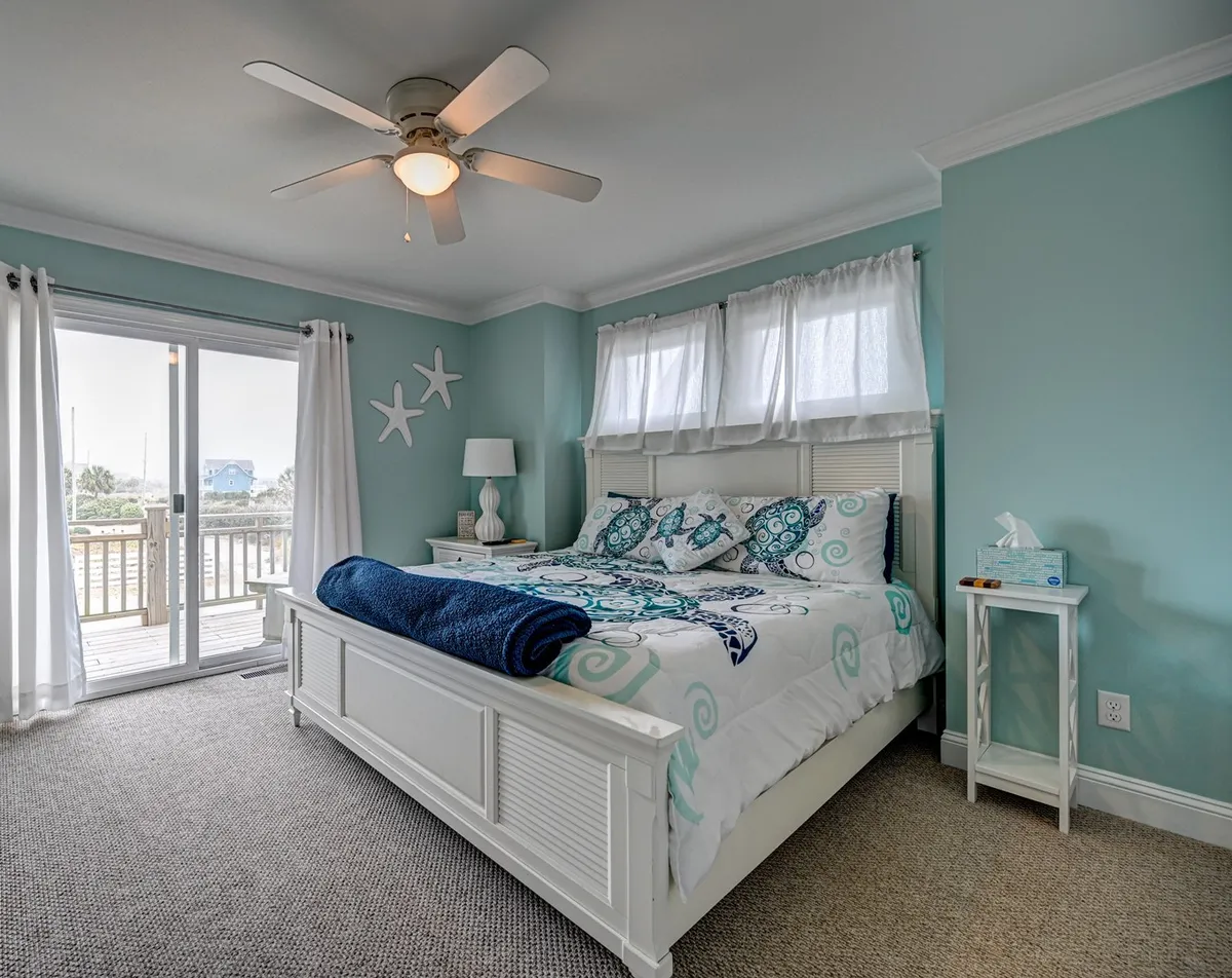 This image showcases a serene king bedroom featuring a comfortable bed, sliding glass doors that open to a deck, decorative starfish accents, and a ceiling fan. The room is filled with natural light and a gentle coastal ambiance, suggesting access to outdoor space and a tranquil atmosphere ideal for restful nights.