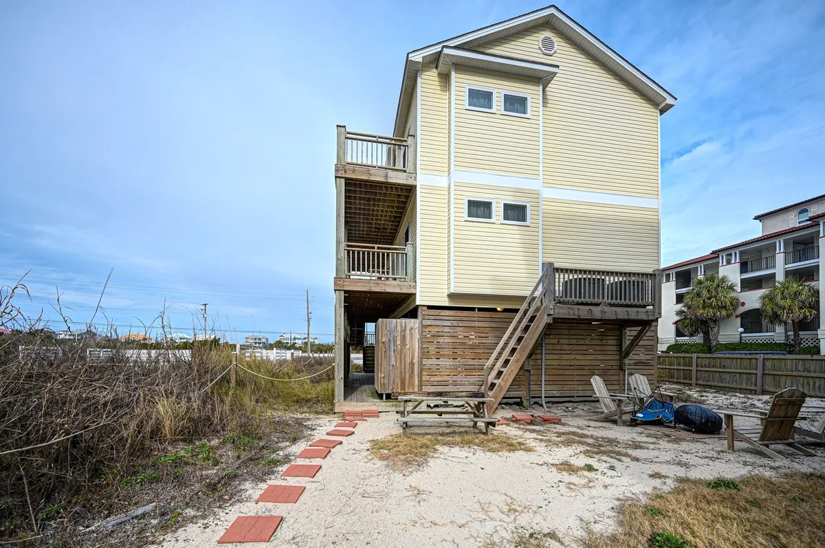 An inviting exterior scene with wooden stairs leading to a deck, a sandy pathway lined with red tiles, and the yellow facade of the house. The proximity to the beach and the deck’s welcoming vibe highlight effortless access to the shore and the home’s beachside charm.