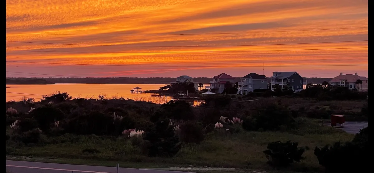 A mesmerizing view of the serene sunset over the water, with soft reflections and silhouettes of houses and lush vegetation. The image captures the peaceful beauty of the area, suggesting moments spent strolling, dining al fresco, or simply savoring the evening light by the shore.