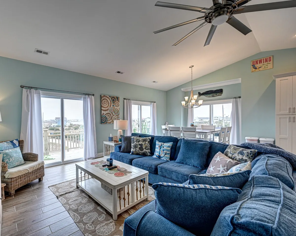 This image showcases a spacious, open-concept great room featuring a large blue sectional sofa, a wooden coffee table, a modern dining area with white chairs, and sliding glass doors leading directly to a sun deck. The room is filled with natural light and offers a seamless flow between the living, dining, and outdoor areas, emphasizing the coastal atmosphere.