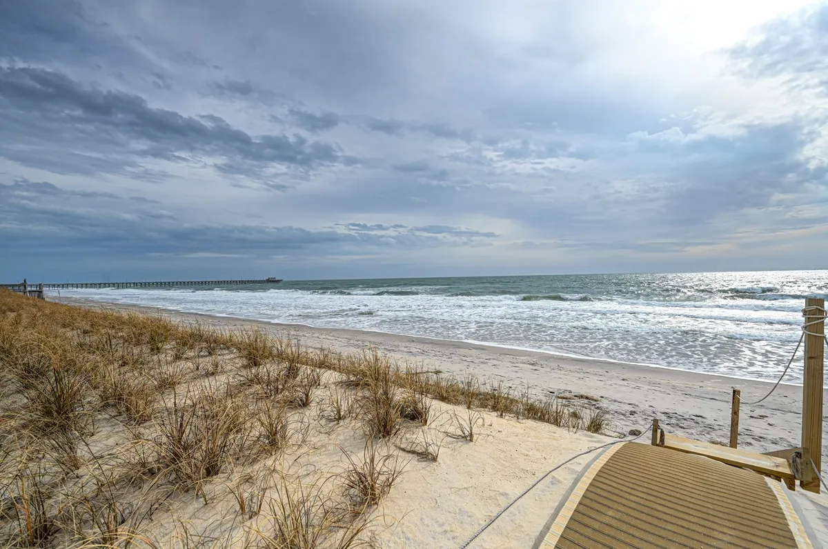 A sweeping view of North Topsail Beach, with gentle ocean waves meeting the sandy shore. Dune grasses line the path, while a pier stretches out in the distance. The scene embodies the quintessential beach experience: open skies, sparkling water, and a vast expanse of sand perfect for family adventures.